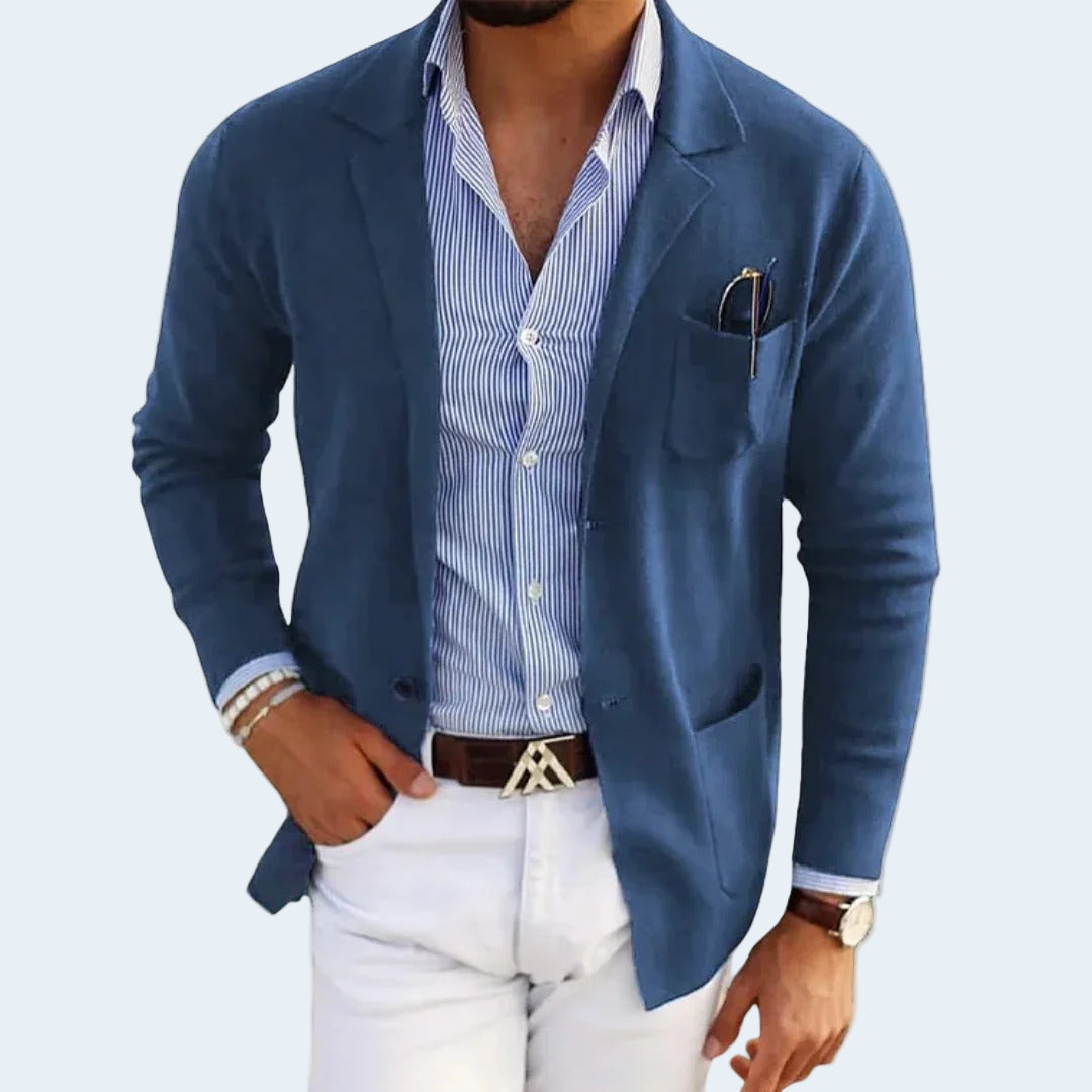 Man wearing a blue blazer over a striped shirt with white pants on a light gray background