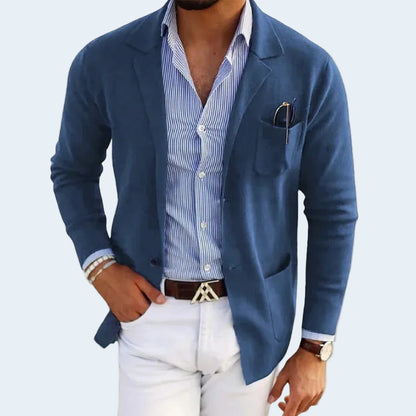 Man wearing a blue blazer over a striped shirt with white pants on a light gray background
