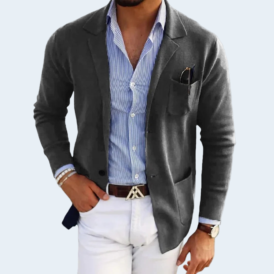 Man wearing a gray blazer over a blue striped shirt with white pants on a light gray background