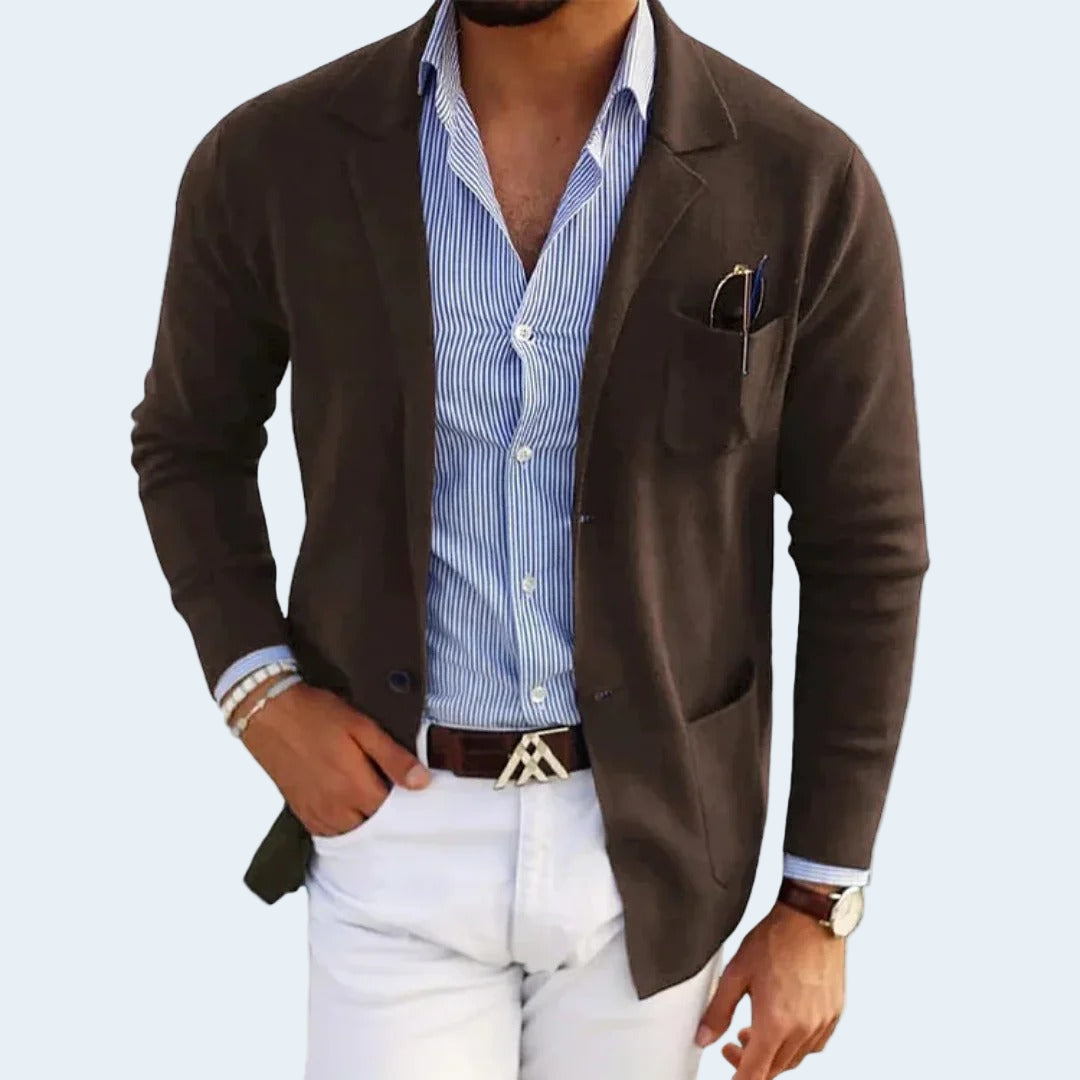 Man wearing a brown blazer over a blue striped shirt with white pants on a light gray background