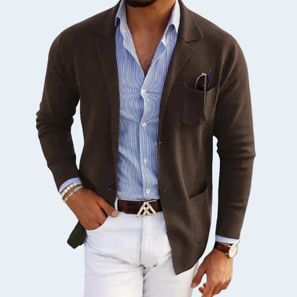 Man wearing a brown blazer over a blue striped shirt with white pants on a light gray background