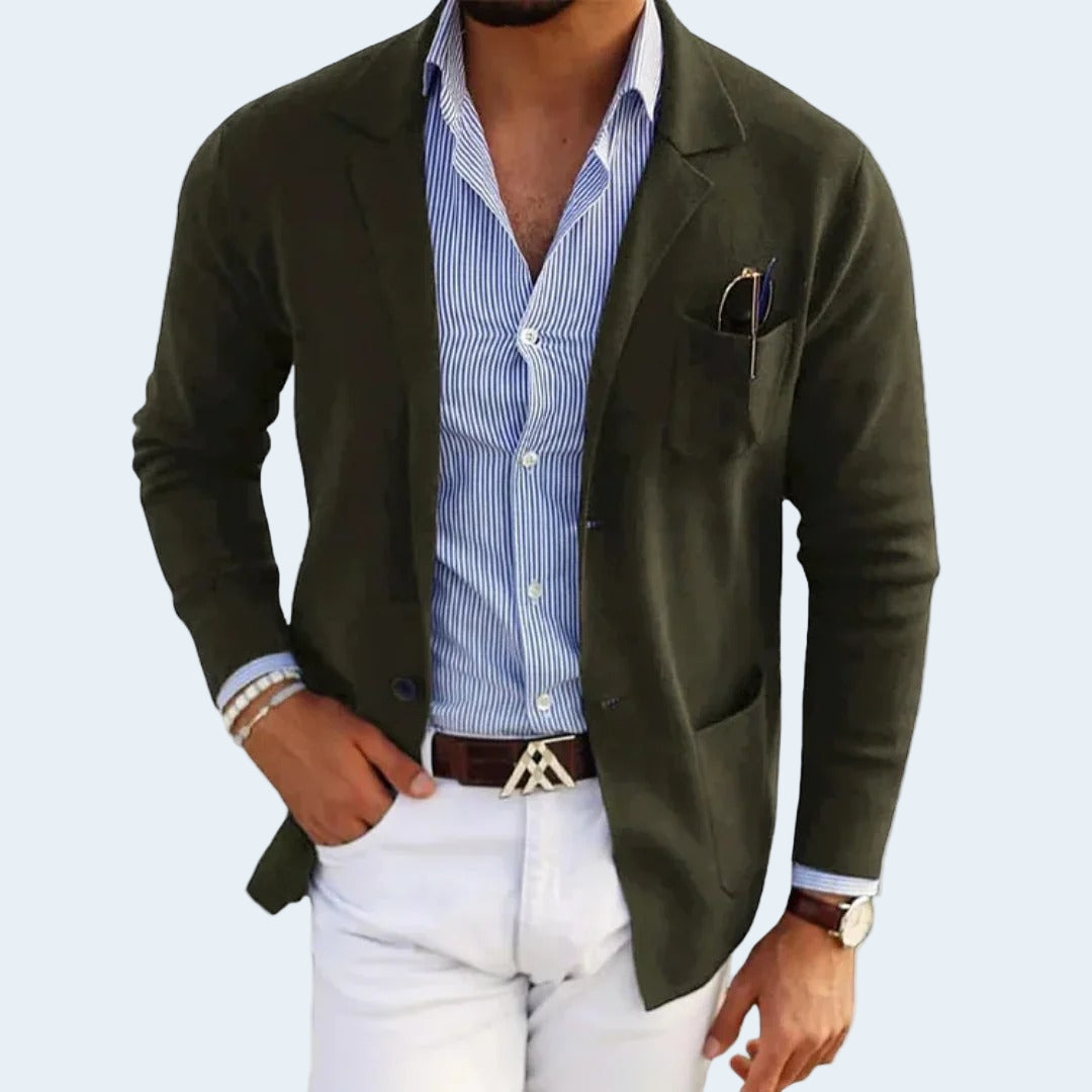 Man wearing a green blazer over a blue striped shirt with white pants on a light gray background