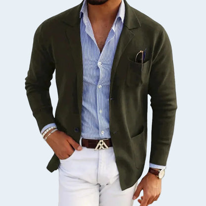 Man wearing a green blazer over a blue striped shirt with white pants on a light gray background